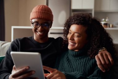 Modern apartmandaki koltukta oturmuş tablete gülümseyen çok ırklı, modaya uygun bir çift.