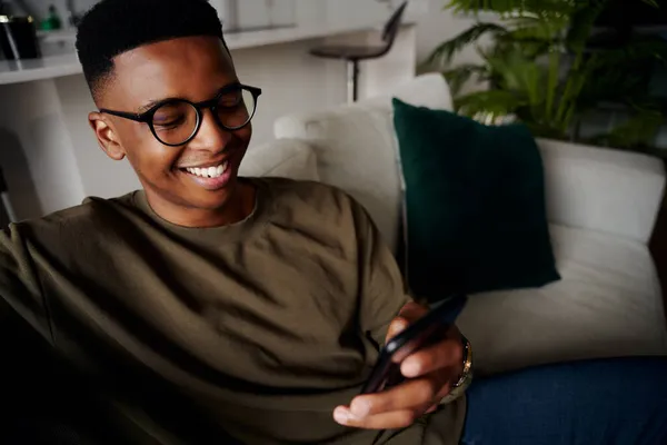 Joven adulto negro masculino relajado, sonriendo mientras envía mensajes de texto en el teléfono inteligente en el sofá — Foto de Stock