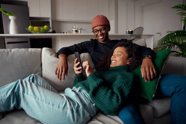 Багатоетнічна пара сміється, дивлячись на текст по телефону під час перебування на дивані в сучасній квартирі — стокове фото