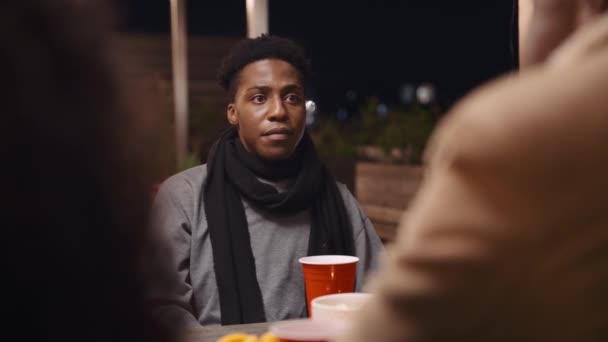 Hombre adulto negro sentado en una mesa en discusión con amigos en una fiesta en la azotea en la noche. — Vídeo de stock