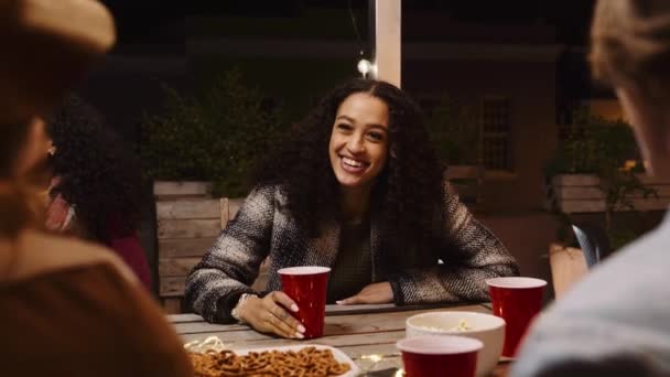 Hembra multicultural charlando, asintiendo y sentada en una mesa en una fiesta en la azotea de la ciudad durante la noche — Vídeos de Stock
