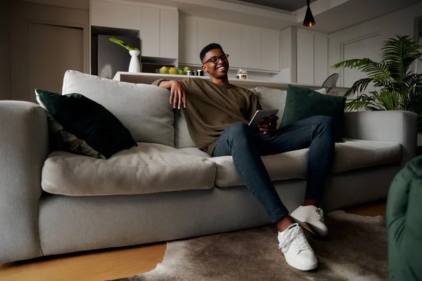 Молодий дорослий чорний чоловік посміхається на дивані під час використання планшета, сучасна квартира — стокове фото