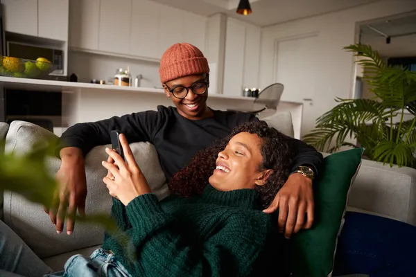 Multi-ethnic couple looking at each other, laughing about text on phone. On sofa in modern apartment — Stock Photo, Image