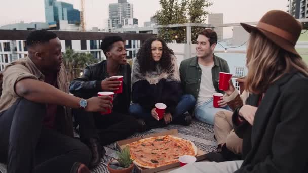 Grupo multiétnico de amigos adultos sentados, comendo pizza e dizendo saúde em um telhado na luz da noite. Imagens de alta qualidade — Vídeo de Stock
