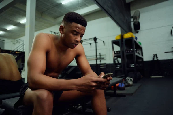 Молодий афроамериканець, який дивиться на телефон, щоб відповісти на повідомлення в Інтернеті. Чоловік, особистий тренер, дивиться на телефон, сидячи в спортзалі. Фотографія високої якості — стокове фото