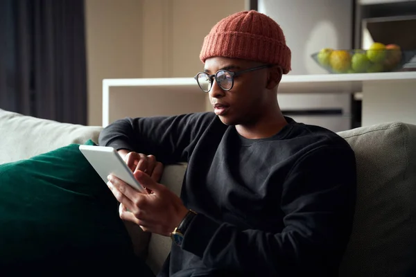 Adulto macho preto concentrando-se enquanto olha para tablet. Apartamento moderno no sofá — Fotografia de Stock
