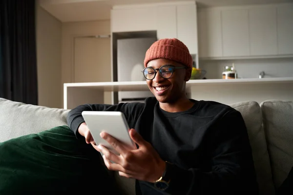 Взрослый черный мужчина улыбается, глядя на планшет. Современная квартира на диване — стоковое фото