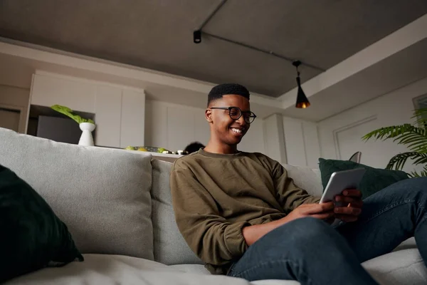 행복 한 젊은 흑인 남성 소파에서 태블릿, 현대 아파트를 사용하는 동안 — 스톡 사진
