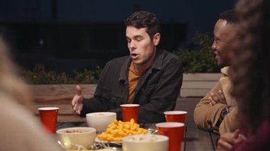 Beyaz erkek, şehirdeki bir çatı partisinde masanın etrafında çeşitli arkadaşlarıyla sosyalleşiyor.
