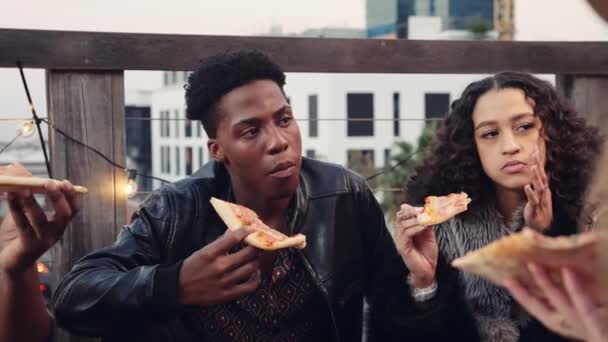 Hombre negro socializando, comiendo pizza y charlando con diversos grupos de amigos adultos en una azotea al atardecer en la ciudad — Vídeo de stock