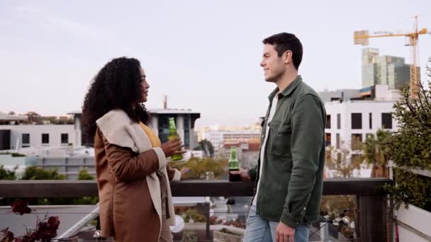 Beyaz erkek ve çok kültürlü bir kadın alacakaranlıkta şehrin çatısında içkilerle sohbet ediyorlar. — Stok video