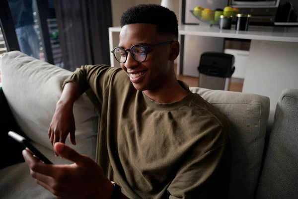 Jeune homme noir adulte avec des lunettes, souriant tout en envoyant des SMS sur smartphone sur le canapé — Photo