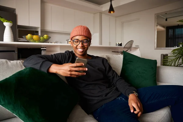 Adult black male smiling while looking at cellphone. Modern apartment on the sofa — Stock Photo, Image