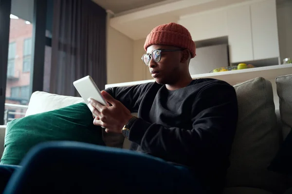 Baixo ângulo de concentração do macho preto adulto enquanto olha para o tablet. Apartamento moderno no sofá — Fotografia de Stock