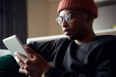 Yetişkin siyah erkek tablete bakarken konsantre oluyor. Kanepede modern daire.