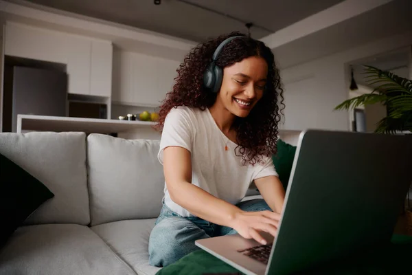 Multi-cultural jovem adulto feminino digitando em seu laptop sentado no sofá enquanto ouve música — Fotografia de Stock