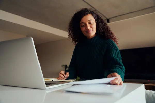 Volwassen multi-culturele vrouw glimlachend verwijzend naar notities tijdens het werken op laptop zittend aan aanrecht top — Stockfoto