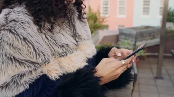 Multi-cultural female texting on smartphone on a rooftop in the city at dusk. Colorful cityscape in background — Stock Video