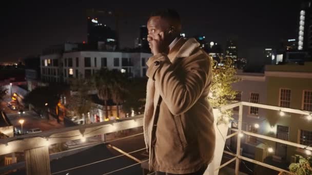 Zwarte man maakt een telefoontje op een dak op een feest met de stadsverlichting op de achtergrond — Stockvideo