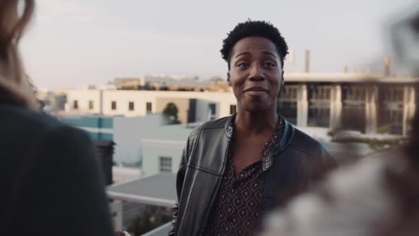 Black male socialising with a diverse group of adult friends at rooftop party. Chatting lightheartedly at dusk. High quality image. — Stock Video