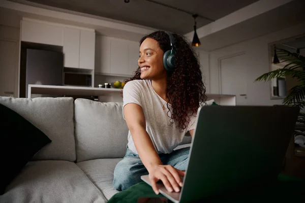 Mulher multicultural sorrindo fora da tela enquanto estava em seu laptop no sofá, ouvindo música — Fotografia de Stock