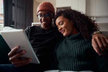 Gülümseyen yetişkin bir çift, çok ırklı, tablet kullanarak modern apartmandaki koltukta yan yana oturuyorlar.