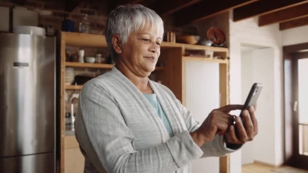 Mensajería de anciana multiétnica jubilada en el teléfono celular. De pie en la cocina moderna, feliz — Vídeos de Stock