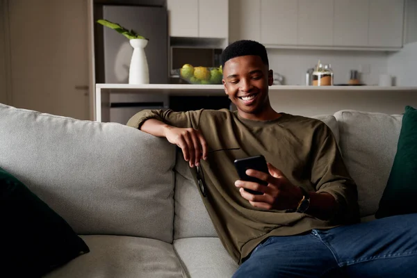 Junge erwachsene schwarze Männchen entspannen sich auf dem Sofa, während sie in einer modernen Wohnung SMS schreiben — Stockfoto