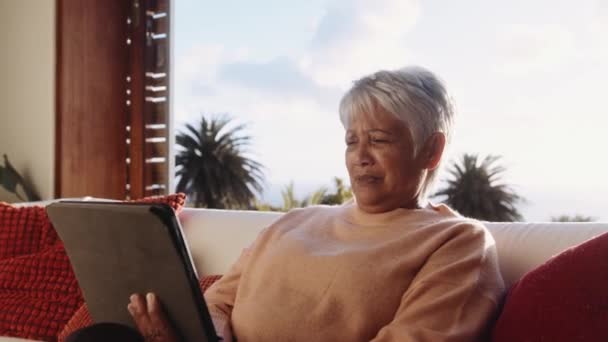 Mujer anciana multicultural sentada en el sofá, saludando a los miembros de la familia llamada en línea utilizando tableta electrónica. — Vídeos de Stock