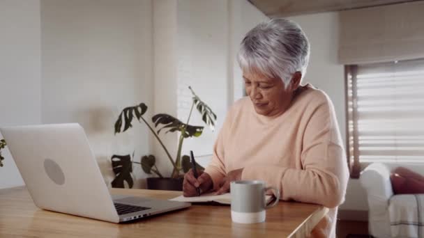 Multi-culturele oudere vrouw scrollen online op laptop, zittend aan de aanrecht. Notities over pen en papier — Stockvideo