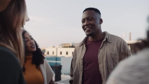 Tall black male socialising with a diverse group of adult friends at rooftop party. Chatting lightheartedly at dusk. High quality image. — Stock Video