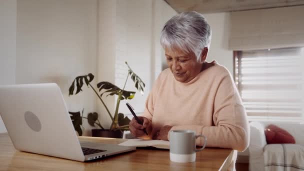 Mujer anciana multicultural que investiga en línea en el ordenador portátil, sentado en el mostrador de la cocina. Notas para la toma de papel y pluma — Vídeos de Stock