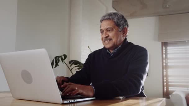 Hombre de edad avanzada multicultural sonriendo, escribiendo en el ordenador portátil, sentado en el mostrador de la cocina — Vídeos de Stock