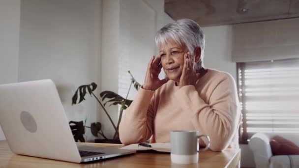 Mujer anciana multicultural interesada, recibiendo malas noticias mientras está en el portátil, sentada en el mostrador de la cocina — Vídeo de stock