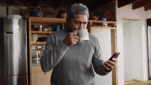 Multi-cultural elderly male scrolling on phone while drinking morning coffee in kitchen — Stock Video