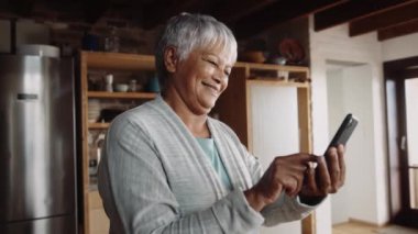 Çok kültürlü, yaşlı ve akıllı telefonuyla yazan emekli bir kadın. Modern mutfakta duruyor.