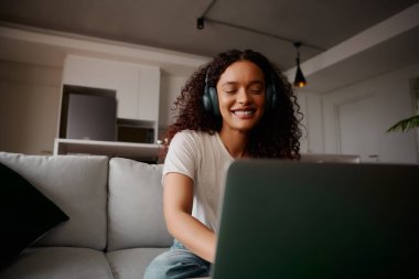 Çok kültürlü bir kadın kanepede oturmuş müzik dinlerken dizüstü bilgisayara gülümsüyor.