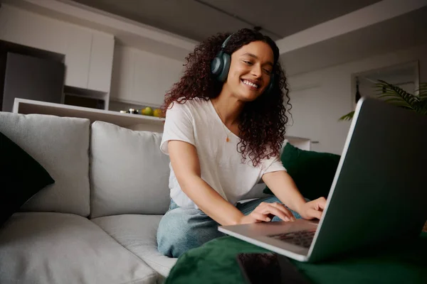 Multi-kulturális nő a laptopján, vidáman ül a kanapén, miközben zenét hallgat — Stock Fotó