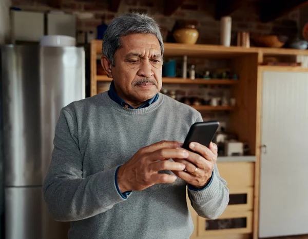 Серьезный многокультурный пожилой мужчина печатает сообщение на смартфоне, стоя на современной кухне. — стоковое фото
