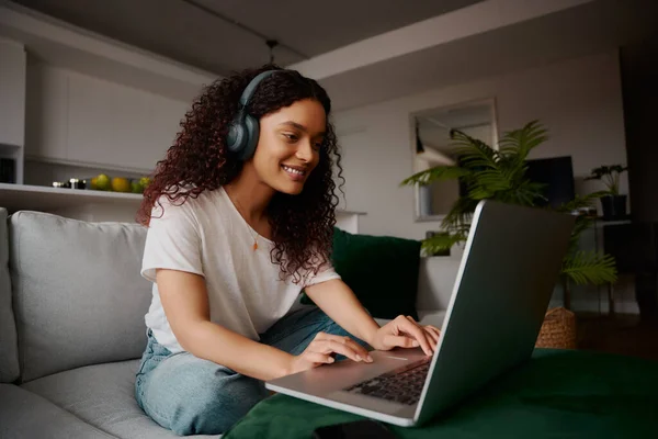 Mulher multicultural digitando em seu laptop sentado no sofá enquanto ouve música — Fotografia de Stock