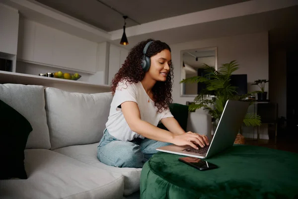 Усміхнена мультикультурна жінка на своєму ноутбуці сидить на дивані з навушниками на — стокове фото