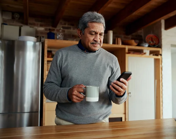 Мультикультурный пожилой мужчина читает ежедневные новости на смартфоне, стоя на современной кухне. — стоковое фото