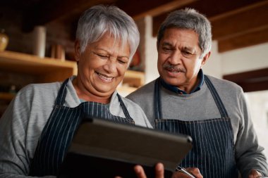 Çok kültürlü yaşlı çift gülüyor, tablet kullanarak modern mutfakta yemek tarifi araştırıyor. Mutlu çift sağlıklı yemek pişiriyor..