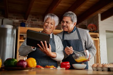 Çok kültürlü, gülümseyen yaşlı bir çift modern mutfakta yemek tarifi için tablet kullanıyor. Mutlu çift sağlıklı yemek pişiriyor..