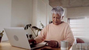 Yaşlı çok kültürlü bir kadın evde dizüstü bilgisayar kullanarak online alışveriş yapıyor, mutfak tezgahında oturuyor.