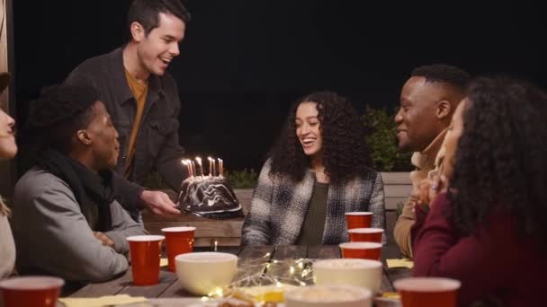 Caucásico macho trayendo pastel de cumpleaños a alegre multi-cultural hembra. Celebrando en una azotea en la noche. — Vídeos de Stock