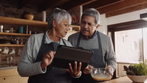 Multikulturní starší pár odkazující na recept na elektronické tabletě. Manžel šlehá vejce v moderní kuchyni — Stock video