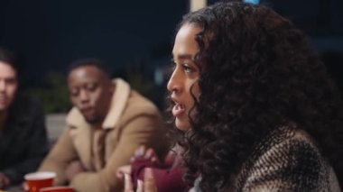 Çok kültürlü bir kadın çeşitli arkadaş grubuyla sohbet ediyor, çatıdaki bir partide masanın etrafında oturuyor..