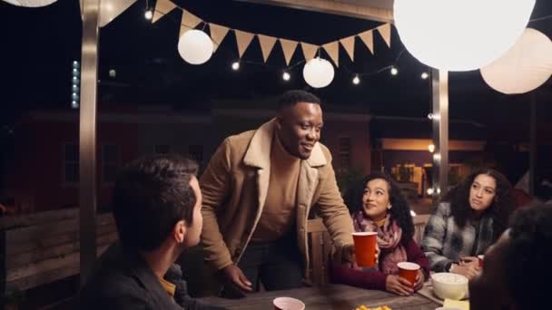 Μαύρος άνδρας κάνει μια πρόποση με την ομάδα των διαφόρων ενηλίκων φίλους του σε ένα πάρτι στην ταράτσα κατά τη διάρκεια της νύχτας — Αρχείο Βίντεο