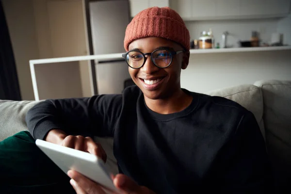 Ein erwachsener schwarzer Mann lächelt in die Kamera, während er ein Tablet benutzt. Moderne Wohnung auf dem Sofa — Stockfoto